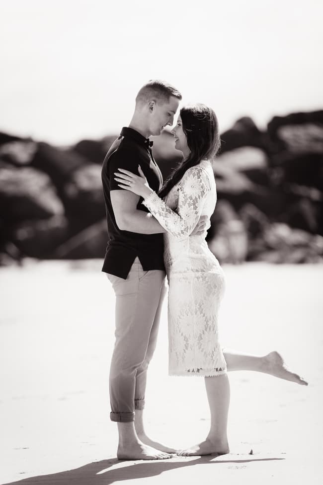 The couple stands close, foreheads touching, on a sandy beach with rocky cliffs in the background.