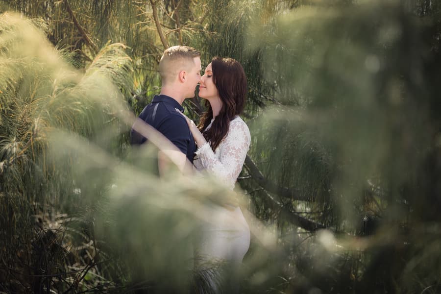 The couple kisses softly, surrounded by lush greenery at The Spit, Gold Coast, with soft natural light.