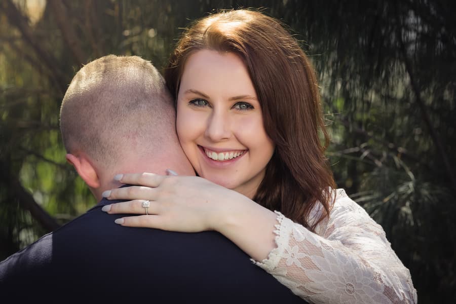 A couple embracing outdoors, with the woman smiling at the camera and showing her engagement ring. Sunlight filters through the trees.