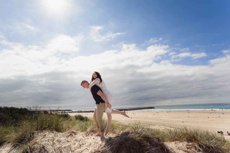 A couple on a sandy dune at The Spit, Gold Coast. The man gives a piggyback ride to the woman under a bright, cloudy sky.
