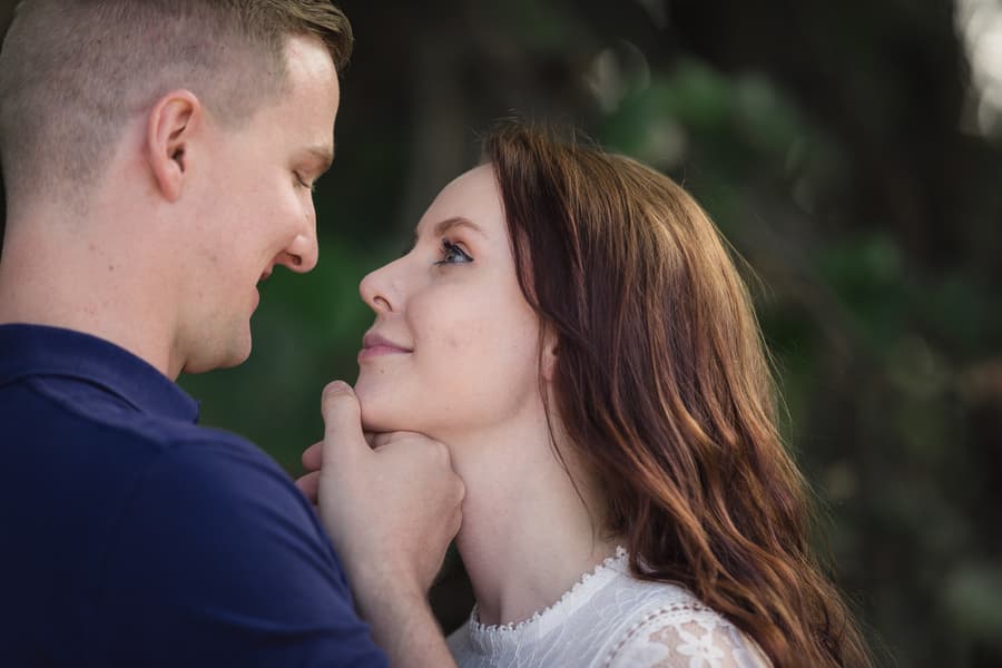 A couple gazing into each other's eyes with a soft, intimate expression. They stand close in a shaded, leafy area.