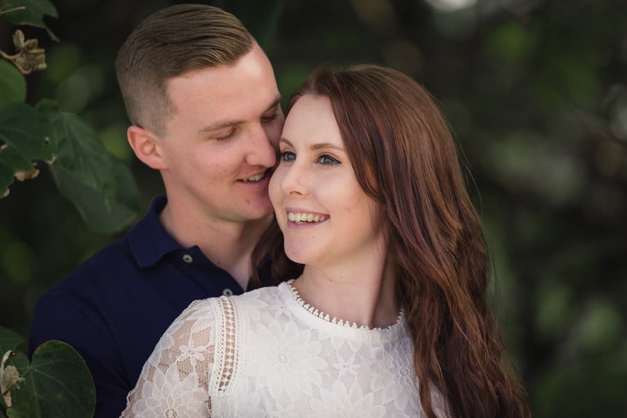 The couple stands closely, smiling and looking away from the camera, surrounded by lush greenery.