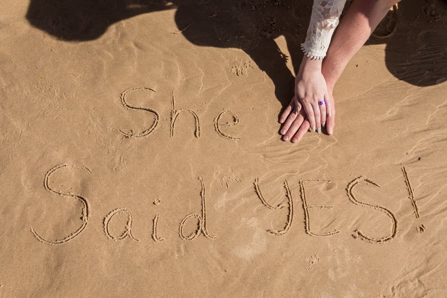 The words 'She Said YES!' written in the sand, with hands gently placed beside the text.