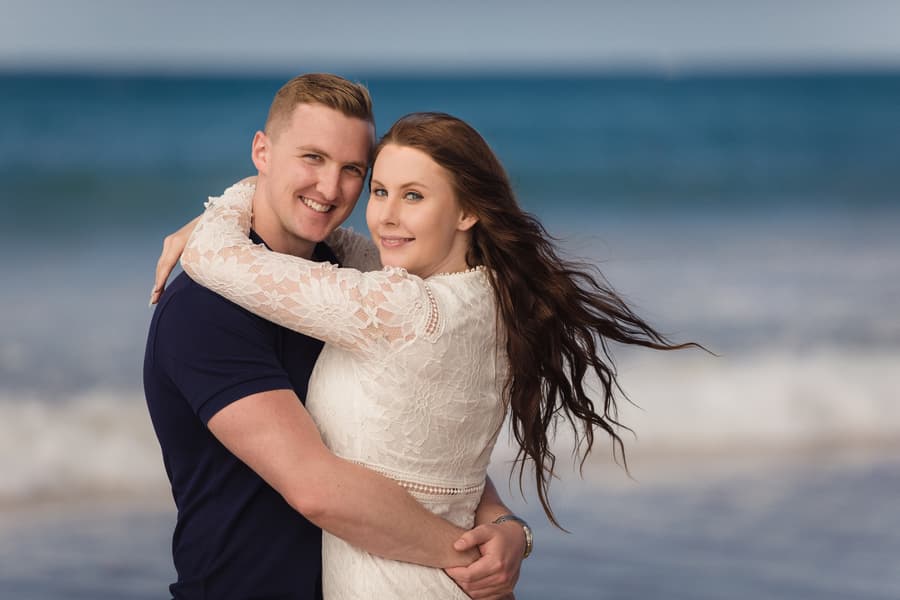 The couple hugging on the beach, smiling at the camera with the ocean in the background.