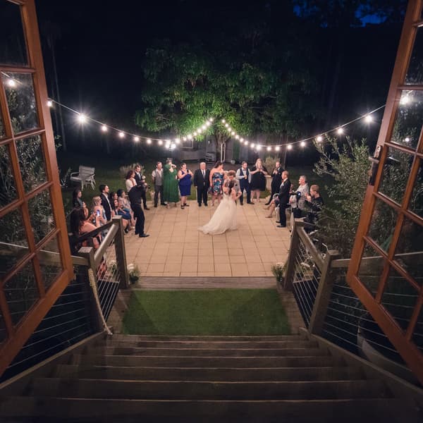 The bride Holly and groom Paul dance together on an outdoor patio at Kwila Lodge, surrounded by guests standing and sitting in a semicircle under string lights at night.