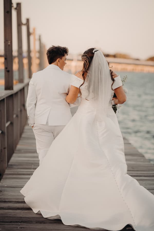Brooke and Tiffany walk arm in arm along a wooden pier at Sandstone Point Hotel, with water visible to the right and a bridge in the background.