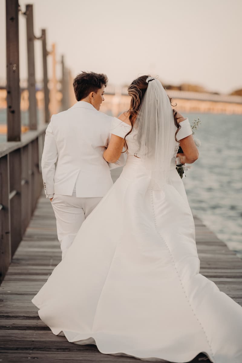 Brooke and Tiffany walk arm in arm along a wooden pier, with water visible to the right and a bridge in the background. Cheap photo booth hire Brisbane by She Said Yes.