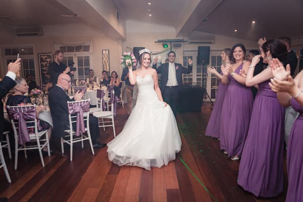 The bride and groom enter the reception at Hillstone St Lucia — The Rosewood Room, with the bride holding her bouquet and bridesmaids in purple dresses clapping on the right, while guests seated at tables applaud on the left.