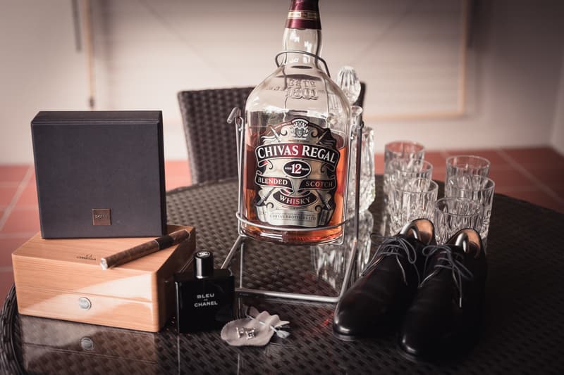 A bottle of Chivas Regal 12-year-old blended Scotch whisky, a set of six crystal glasses, a pair of black dress shoes, a black box with a cigar on top, a wooden box, a bottle of Bleu Chanel cologne, and a small pouch with cufflinks arranged on a round glass table.