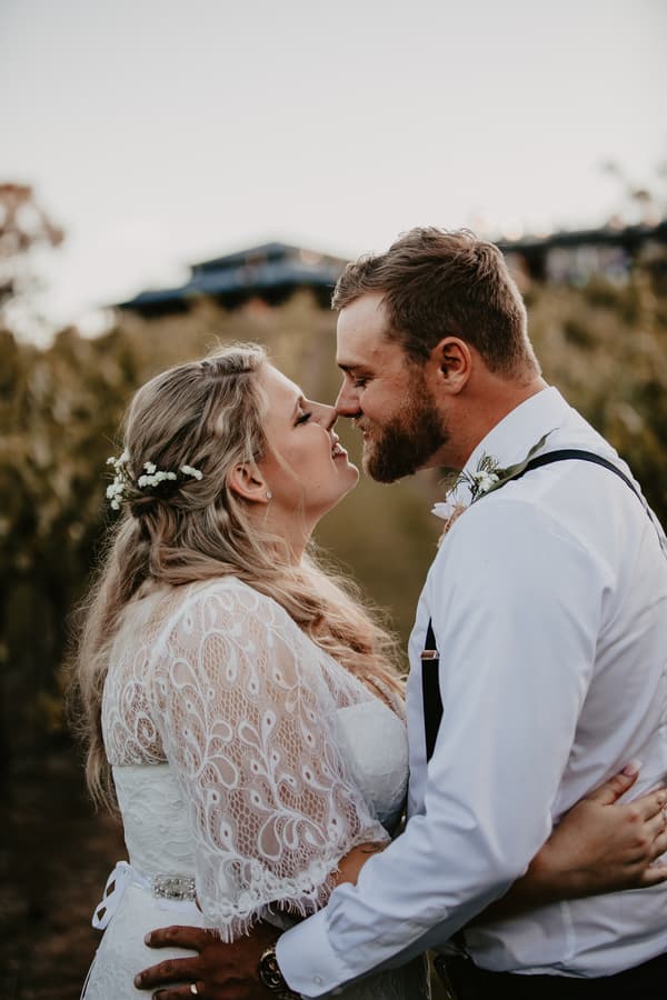 Annie and Trent embrace closely outdoors at Ocean View Estates, with Annie wearing a lace wedding dress and floral hair accents, and Trent in a white shirt with suspenders and boutonniere.