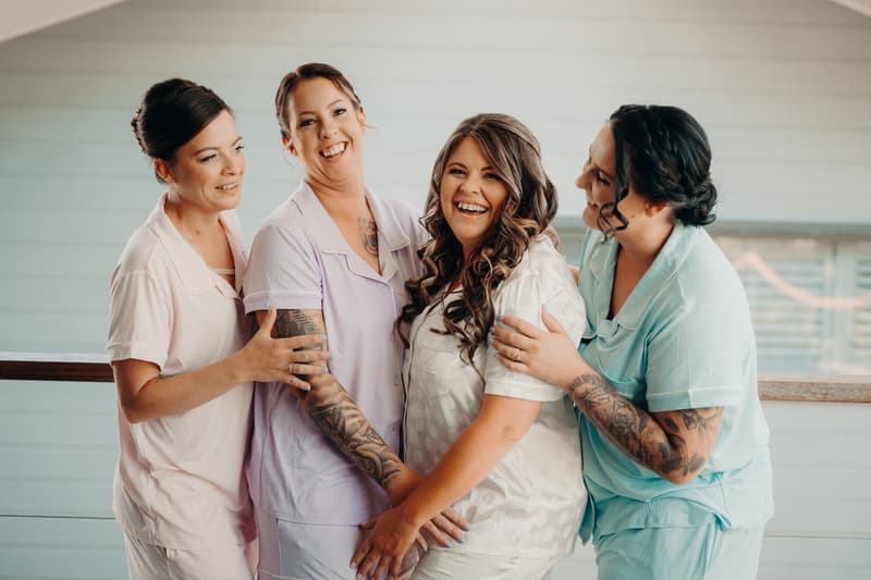 Brooke, the bride, in a white robe is embraced by three women in pastel-colored robes during bridal preparation at Sandstone Point Hotel.
