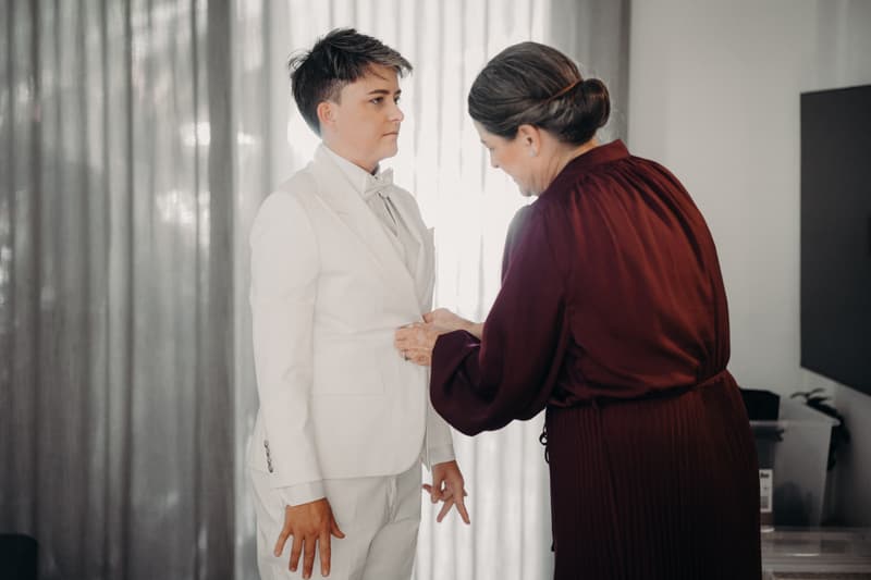 Tiffany, the bride, stands wearing a white suit while an older woman in a burgundy dress adjusts her jacket at Sandstone Point Hotel — The Pavilion.
