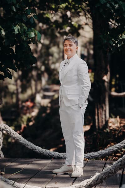 Tiffany, the bride, stands alone outdoors on a wooden platform surrounded by greenery, wearing a white suit with a bow tie and smiling. Wedding live streaming by She Said Yes.