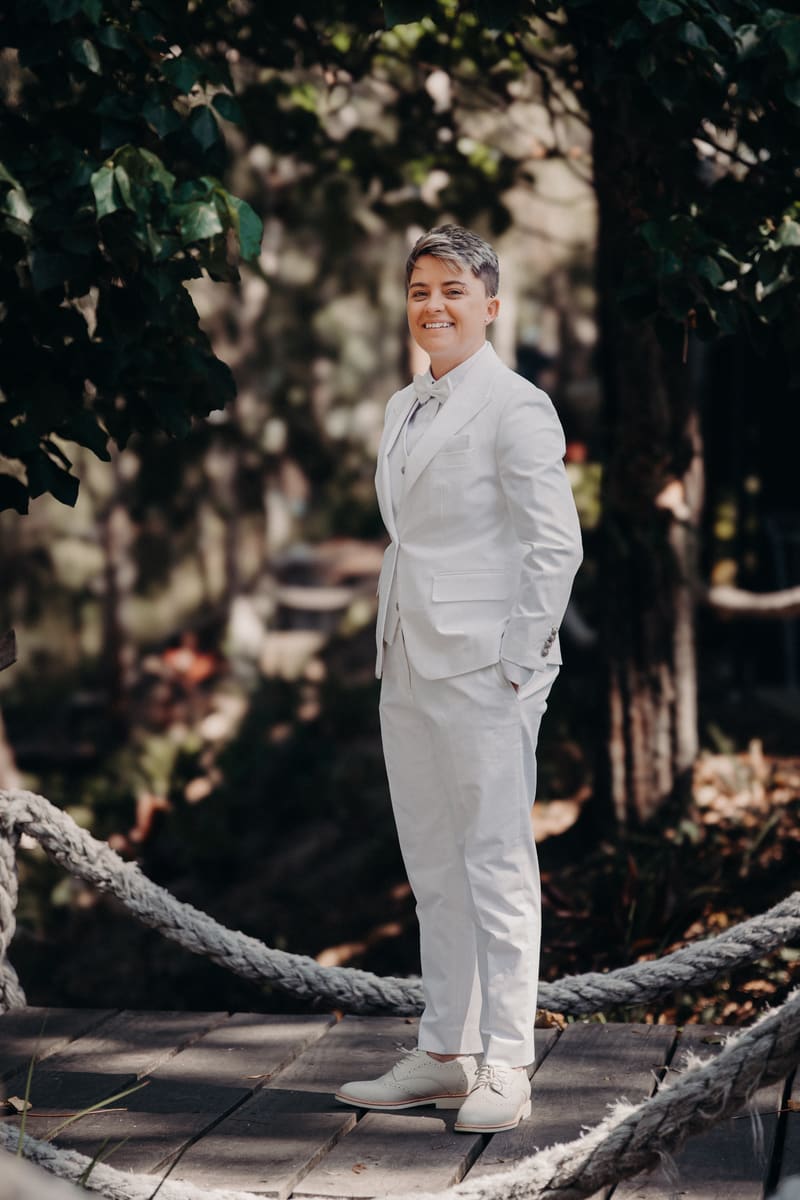 Tiffany, the bride, stands alone outdoors on a wooden platform surrounded by greenery, wearing a white suit with a bow tie and smiling. Winter wedding Brisbane dress code by She Said Yes.
