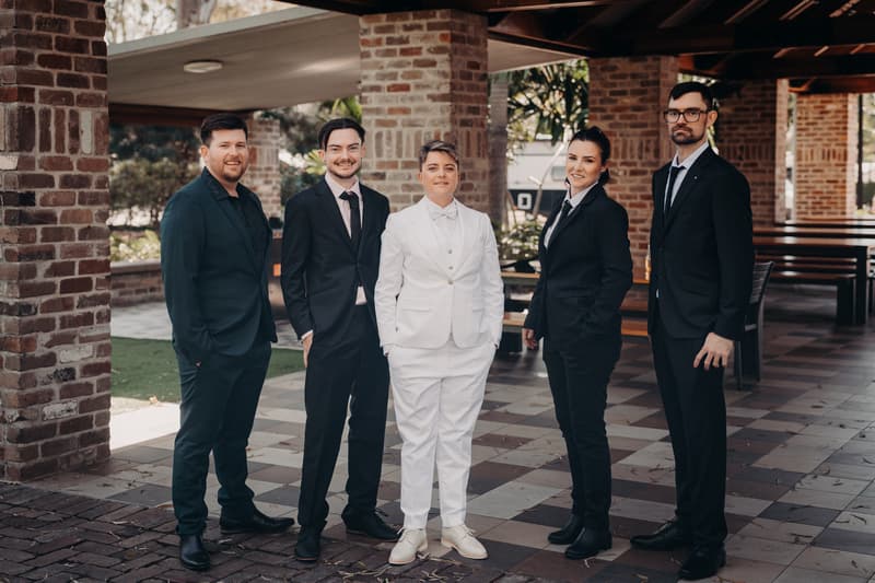 Tiffany in a white suit stands with four wedding party members dressed in black suits under a covered outdoor area at Sandstone Point Hotel — The Pavilion.