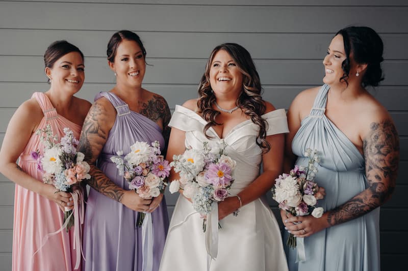 Brooke the bride stands with three bridesmaids holding bouquets against a gray paneled wall at Sandstone Point Hotel — The Pavilion.