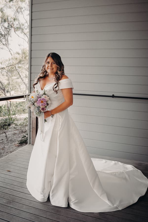 Brooke, the bride, poses alone in her wedding gown holding a bouquet on a wooden deck at Sandstone Point Hotel — The Pavilion.