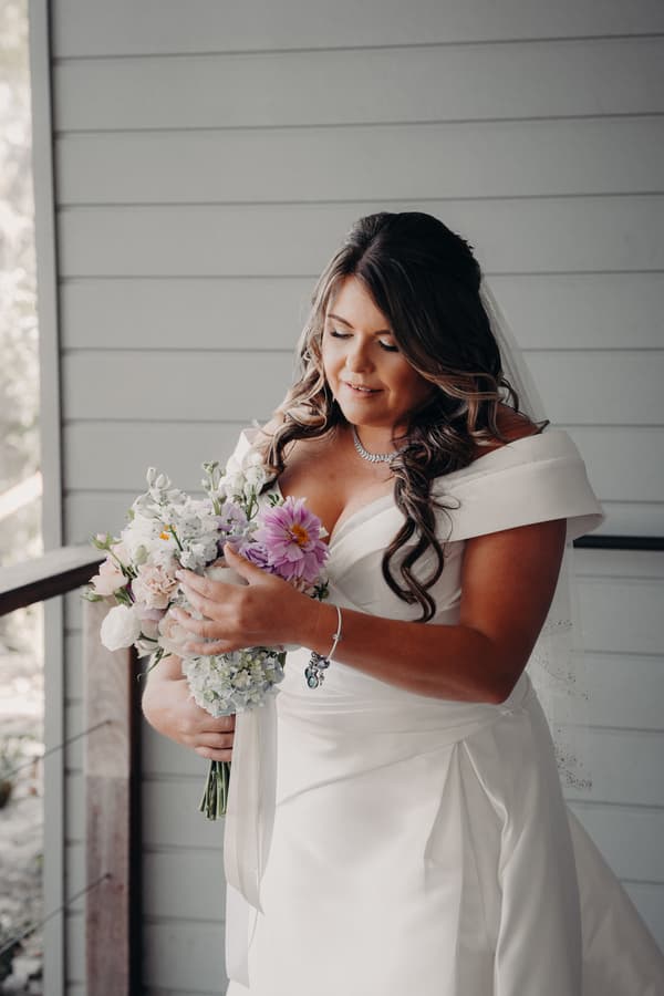 Brooke, the bride, stands holding a bouquet of flowers wearing a white off-shoulder wedding gown and veil at Sandstone Point Hotel — The Pavilion.