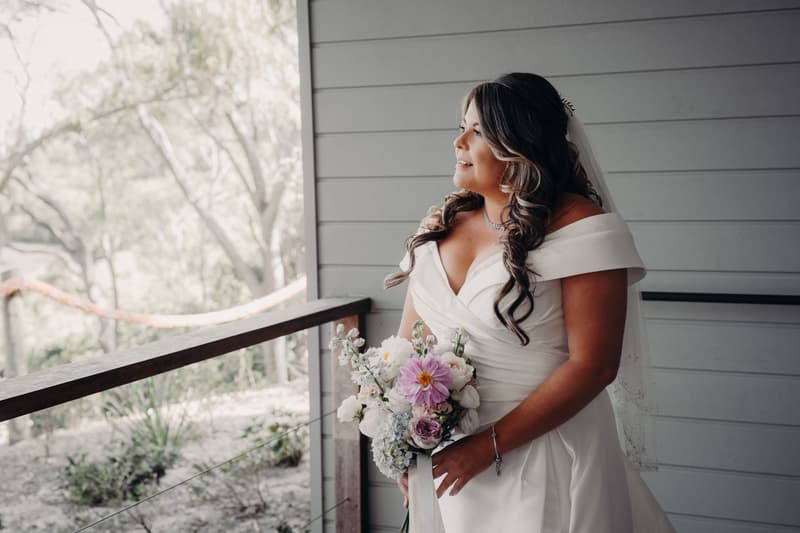 Brooke, the bride, stands holding a bouquet of flowers on a balcony at Sandstone Point Hotel — The Pavilion, looking to the side.