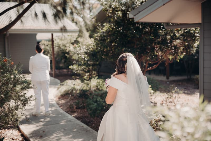 Brooke in a wedding gown approaches Tiffany in a white suit from behind along a garden path at Sandstone Point Hotel.