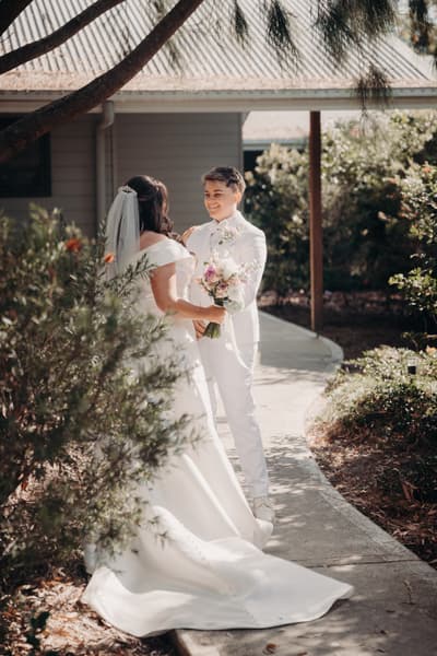 Brooke and Tiffany stand facing each other on a garden path outside a building, The Pavilion. Brooke wears a white wedding gown with a veil and holds a bouquet, while Tiffany wears a white suit and smiles at Brooke. Cinematic wedding photos by She Said Yes.