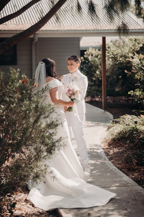 Brooke and Tiffany stand facing each other on a garden path outside a building at Sandstone Point Hotel, The Pavilion. Brooke wears a white wedding gown with a veil and holds a bouquet, while Tiffany wears a white suit and smiles at Brooke.