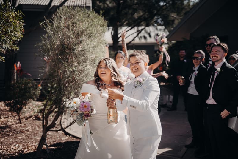 Brooke and Tiffany stand outside at Sandstone Point Hotel — The Pavilion, with Brooke holding a bouquet and Tiffany opening a bottle of sparkling drink, while guests and bridal party members watch and celebrate in the background.