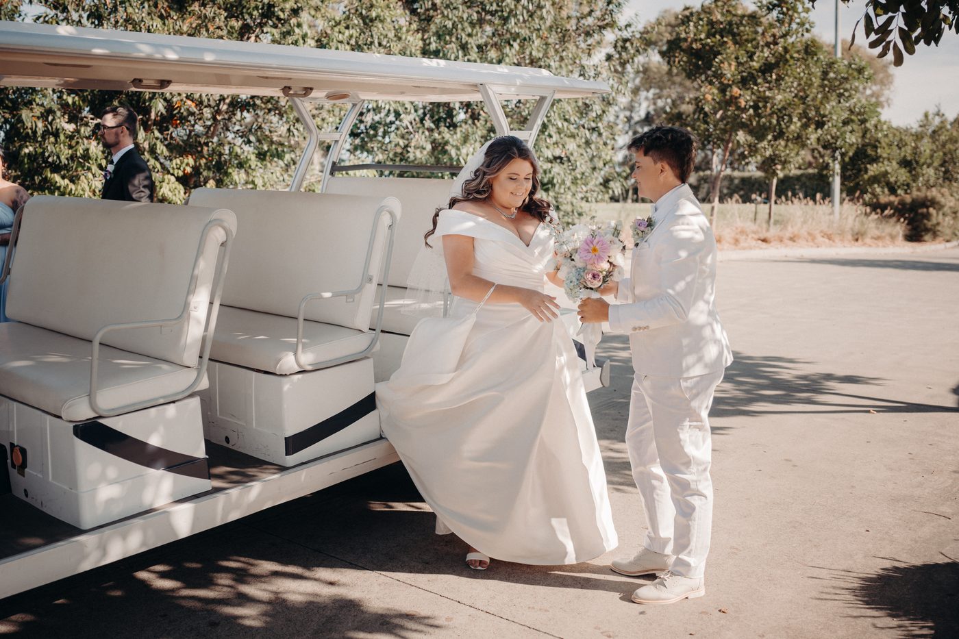 Bride Brooke in a white wedding gown and bride Tiffany in a white suit stand beside a white golf cart, with Brooke holding a bouquet of flowers. Cinematic videographer by She Said Yes.