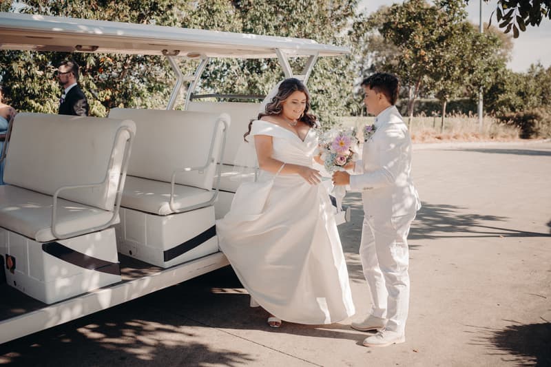 Bride Brooke in a white wedding gown and bride Tiffany in a white suit stand beside a white golf cart at Sandstone Point Hotel — The Pavilion, with Brooke holding a bouquet of flowers.