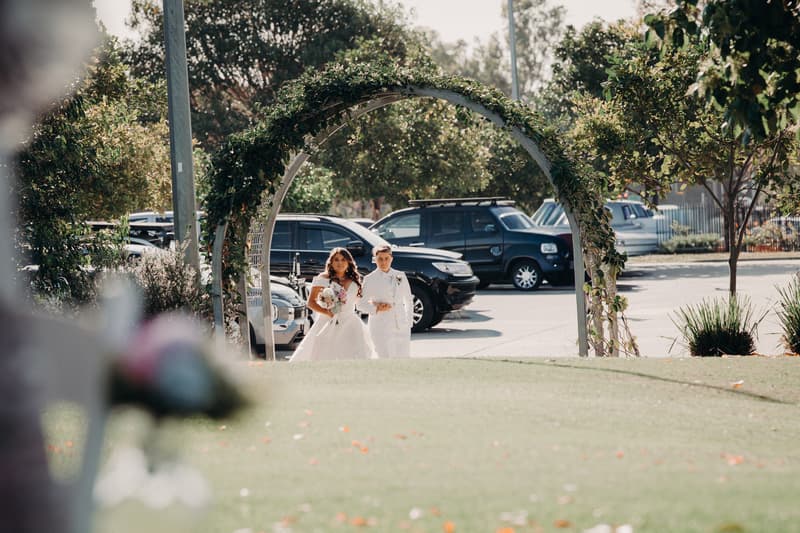 Brides Brooke and Tiffany walk arm in arm under a floral arch at Sandstone Point Hotel — The Pavilion, with parked cars and trees in the background.