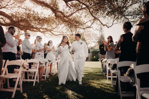 Brides Brooke and Tiffany walk down the aisle arm in arm at Sandstone Point Hotel — The Pavilion, surrounded by applauding guests seated on white chairs decorated with flowers under a large tree.
