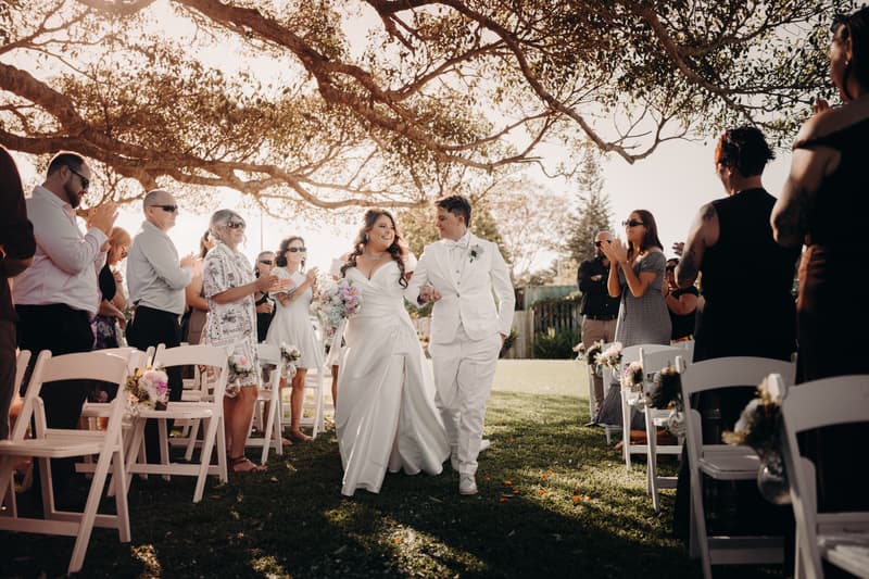 Brides Brooke and Tiffany walk down the aisle arm in arm at Sandstone Point Hotel — The Pavilion, surrounded by applauding guests seated on white chairs decorated with flowers under a large tree.
