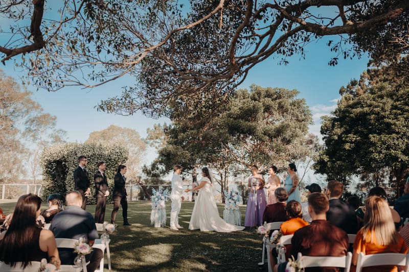 Brides Brooke and Tiffany exchange vows at the ceremony stage, surrounded by bridesmaids in pastel dresses and groomsmen in black suits, with guests seated on white chairs watching. Good wedding vows by She Said Yes.