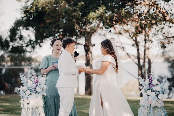 Brides Brooke and Tiffany hold hands facing each other during their wedding ceremony at Sandstone Point Hotel — The Pavilion, with an officiant standing behind them and floral arrangements on either side.