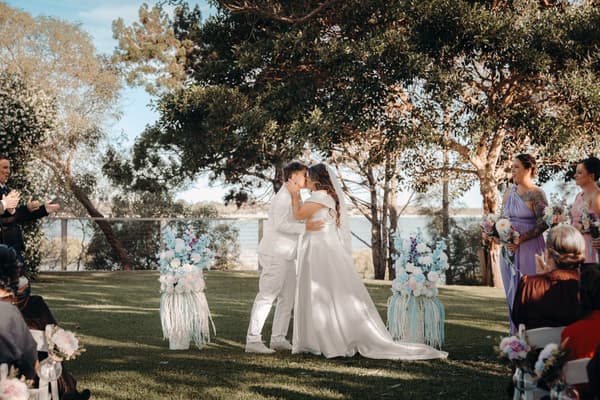 Brides Brooke and Tiffany share a kiss at the ceremony stage at Sandstone Point Hotel — The Pavilion, surrounded by bridesmaids holding bouquets and guests seated on either side.