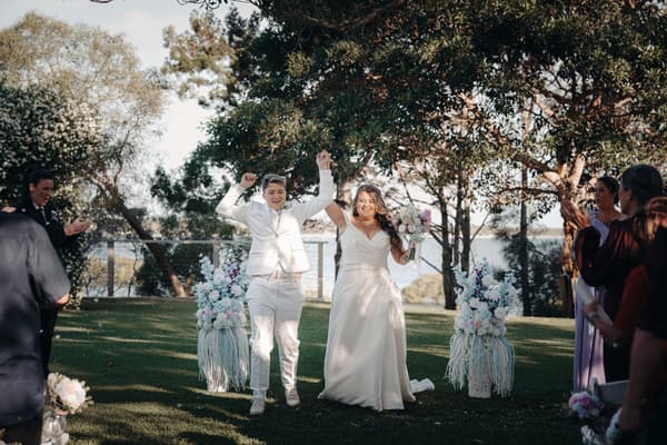 Brides Brooke and Tiffany walk down the aisle together at Sandstone Point Hotel — The Pavilion, holding hands and celebrating as guests applaud on either side.