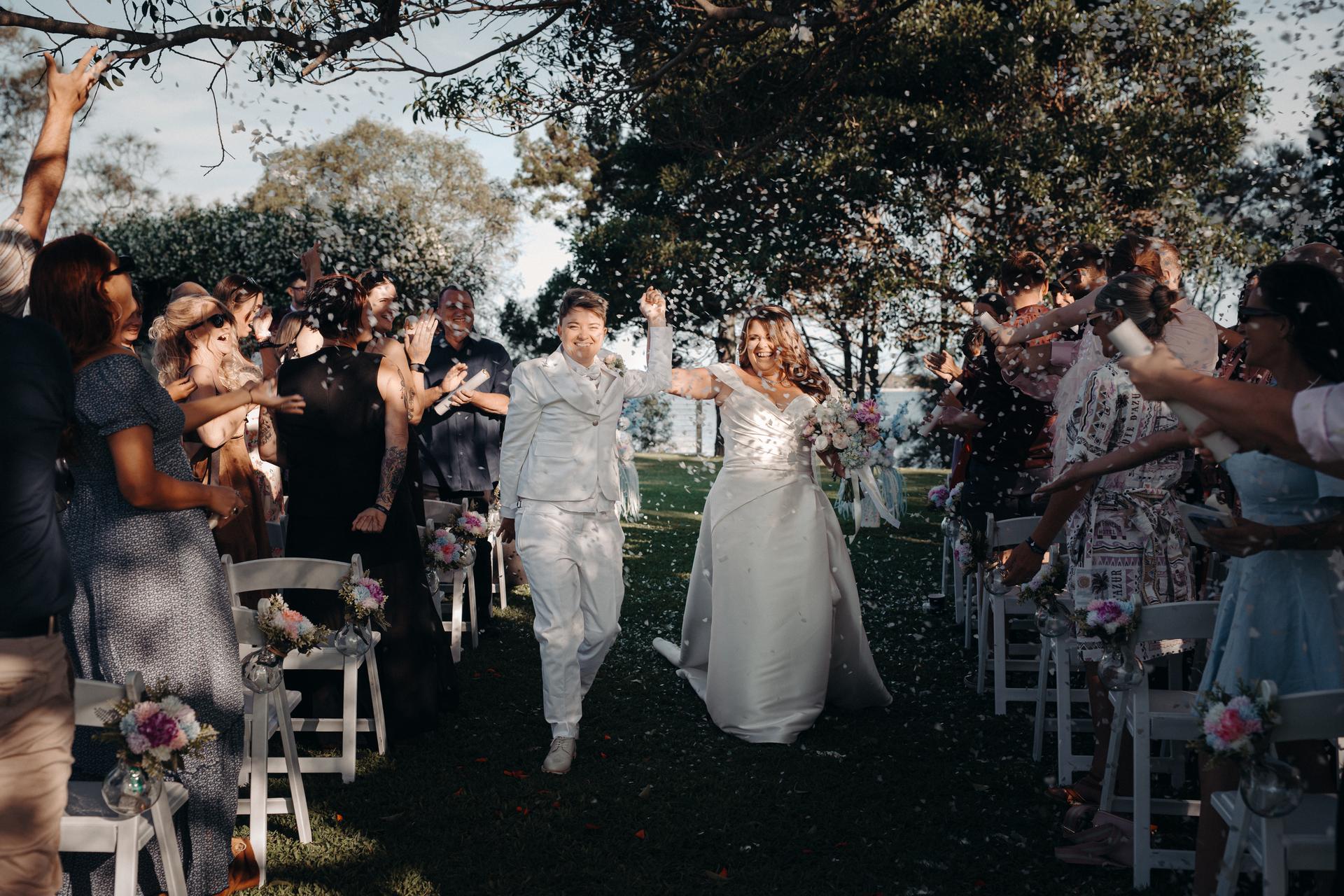 Brides Brooke and Tiffany walk down the aisle together at Sandstone Point Hotel — The Pavilion, with guests on both sides throwing confetti.