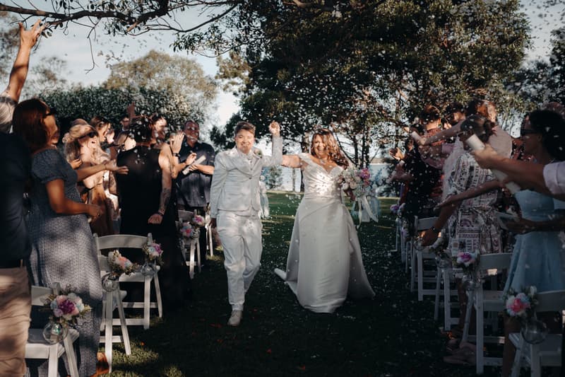 Brides Brooke and Tiffany walk down the aisle together at Sandstone Point Hotel — The Pavilion, with guests on both sides throwing confetti.