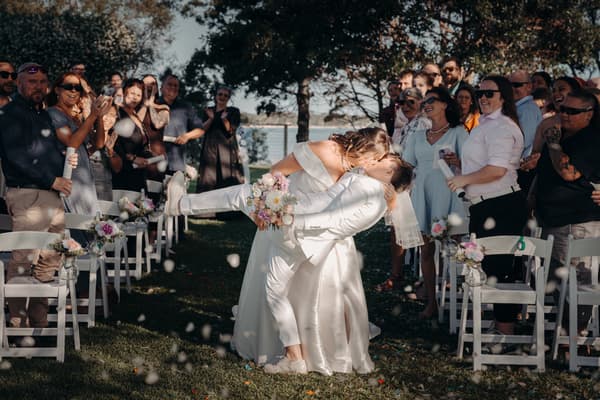 Brooke and Tiffany share a kiss at the Sandstone Point Hotel — The Pavilion ceremony aisle, surrounded by applauding guests seated on white chairs decorated with floral arrangements.