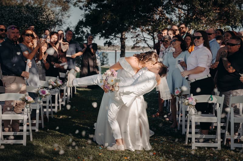 Brooke and Tiffany share a kiss at the Sandstone Point Hotel — The Pavilion ceremony aisle, surrounded by applauding guests seated on white chairs decorated with floral arrangements.