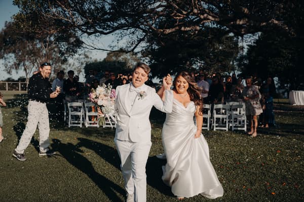 Brooke and Tiffany walk hand in hand down the aisle at Sandstone Point Hotel — The Pavilion after their wedding ceremony, with guests standing and throwing confetti.