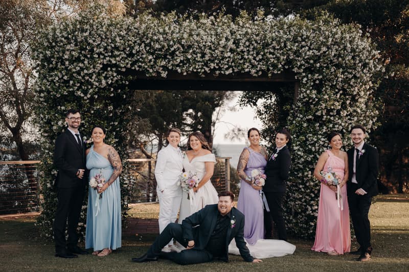 Brooke and Tiffany stand together under a floral arch at Sandstone Point Hotel — The Pavilion, surrounded by their bridal party dressed in pastel gowns and black suits, with one groomsman lying on the grass in front.
