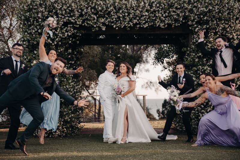 Brooke and Tiffany stand together under a floral arch at Sandstone Point Hotel — The Pavilion, surrounded by their bridal party who are posing playfully.