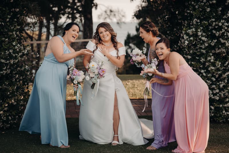 Brooke the bride stands outdoors at Sandstone Point Hotel — The Pavilion with three bridesmaids in pastel dresses holding bouquets, all smiling and interacting playfully.