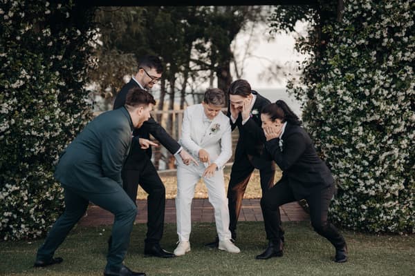 Brooke, the bride in a white suit, shows her wedding ring while surrounded by four members of the bridal party dressed in dark suits. Marriage equality Australia by She Said Yes.