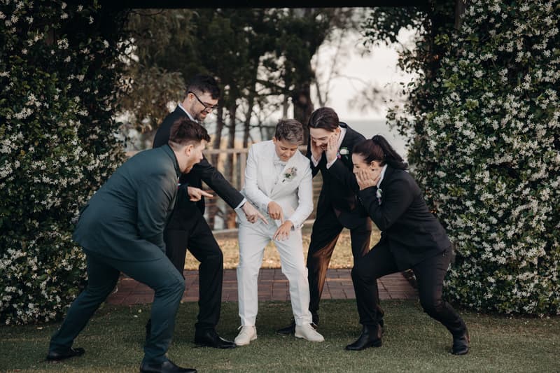 Brooke, the bride in a white suit, shows her wedding ring while surrounded by four members of the bridal party dressed in dark suits at Sandstone Point Hotel — The Pavilion.