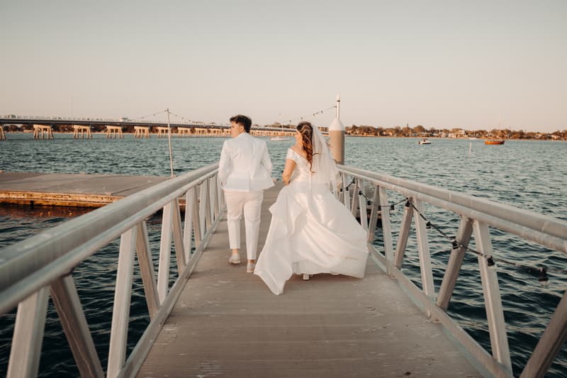 Brooke and Tiffany walk side by side on a pier over water, with Brooke wearing a white wedding gown and veil, and Tiffany dressed in a white suit.