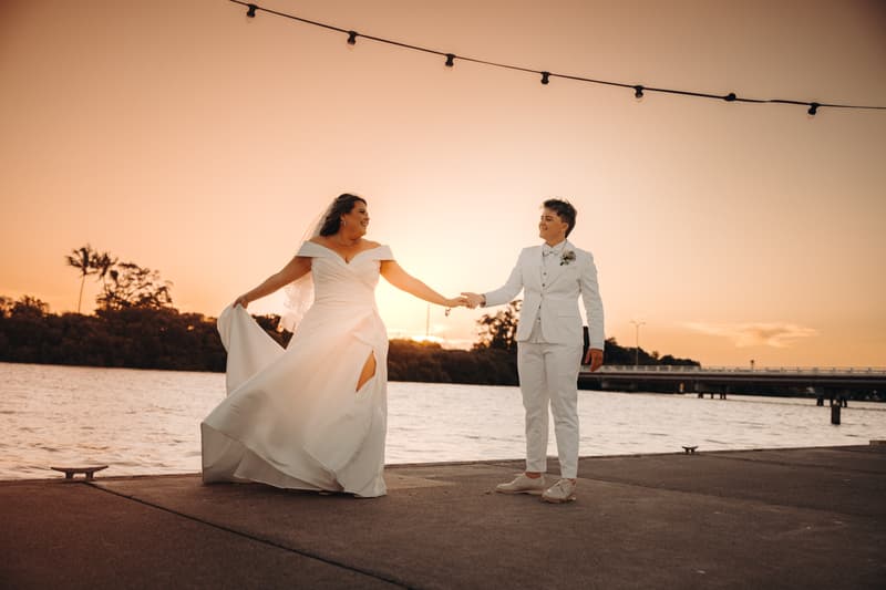 Brooke and Tiffany hold hands and smile at each other near the water at sunset, with Brooke wearing a white wedding gown and Tiffany in a white suit.