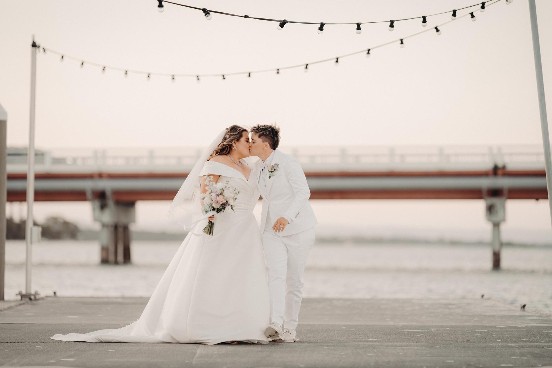 Brooke and Tiffany kiss on a pier with a bridge in the background, Brooke holding a bouquet and wearing a wedding gown, Tiffany in a white suit.