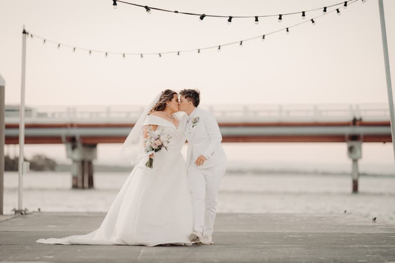 Brooke and Tiffany kiss on a pier with a bridge in the background, Brooke holding a bouquet and wearing a wedding gown, Tiffany in a white suit.